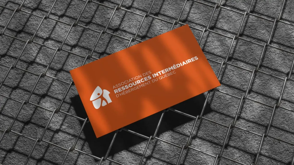 Maquette d'une carte de visite orange avec le logo blanc de l' "Association des Ressources Intermédiaires d'Hébergement du Québec", posée en diagonale sur un grillage métallique gris.