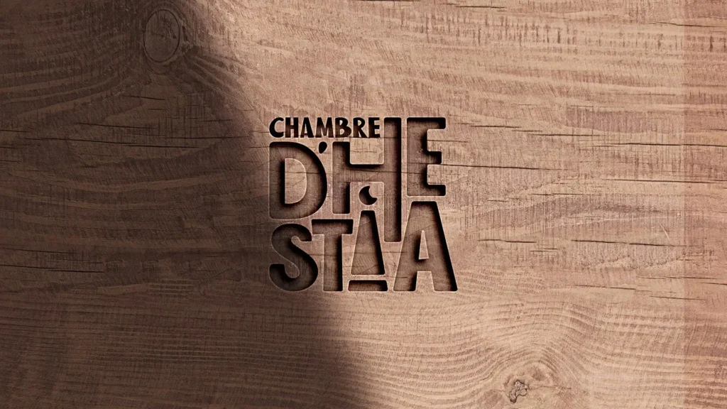 Image montrant le logo "Chambre d'Hestia" gravé en creux dans une surface en bois avec une texture de veinage prononcée et une ombre projetée.