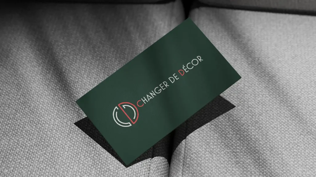 Une carte de visite verte avec le logo "Changer de Décor" en blanc et rouge, posée en diagonale sur un fond gris texturé imitant un tissu.