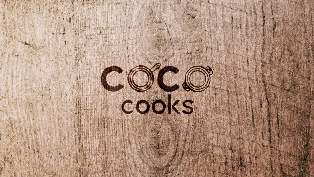 Image montrant le logo "CoCo Cooks" gravé en creux sur une planche de bois avec une texture de veinage visible, avec les "O" stylisés en forme de casseroles.