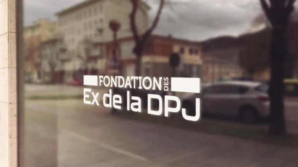 Image d'une vitrine de commerce avec l'inscription blanche "FONDATION DES Ex de la DPJ", reflétant une rue urbaine avec des bâtiments et une voiture.