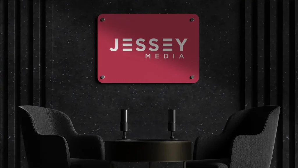 Un panneau carré rouge avec le logo argenté "JESSEY MEDIA", fixé sur un mur texturé noir avec des lamelles verticales, au-dessus de deux fauteuils noirs et une petite table ronde.