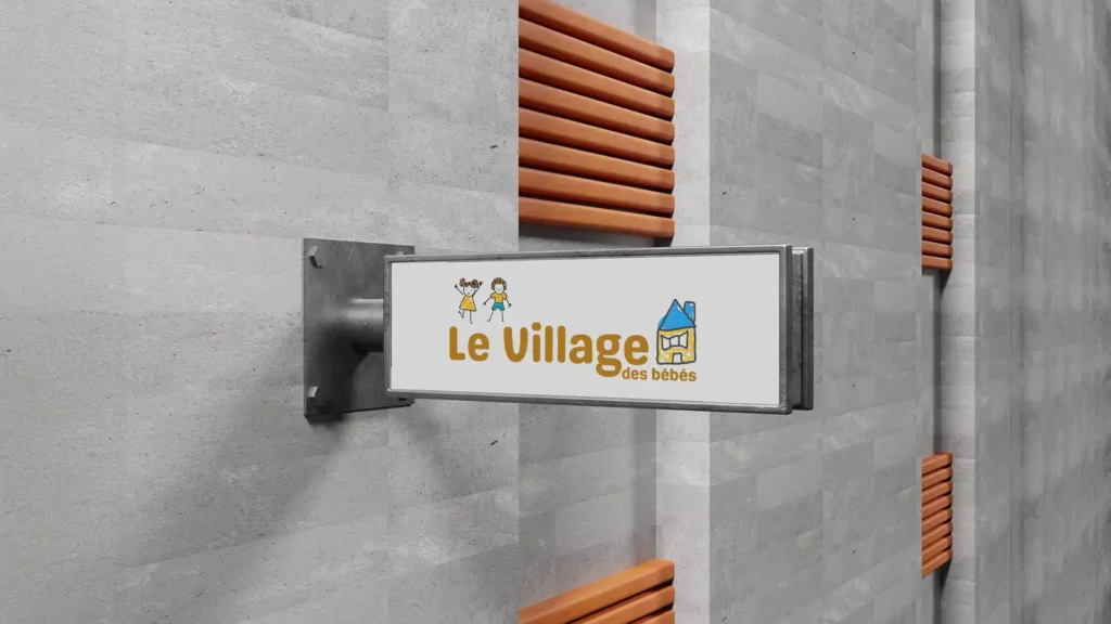 Une enseigne rectangulaire blanche avec le logo "Le Village des bébés" et des illustrations colorées, fixée sur un mur texturé gris avec des éléments décoratifs en bois.