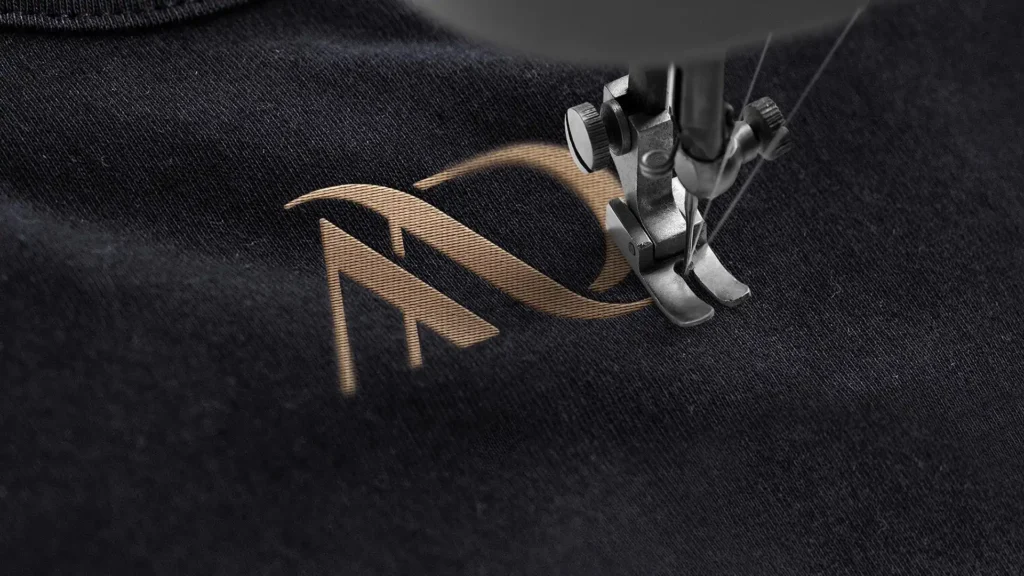 Gros plan d'une machine à coudre brodant un logo doré 'MD' sur un tissu noir.