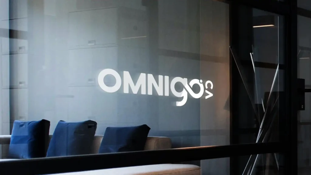 Logo 'OMNIGOS' blanc sur une vitre transparente d'un bureau moderne, avec des coussins bleus visibles en arrière-plan.