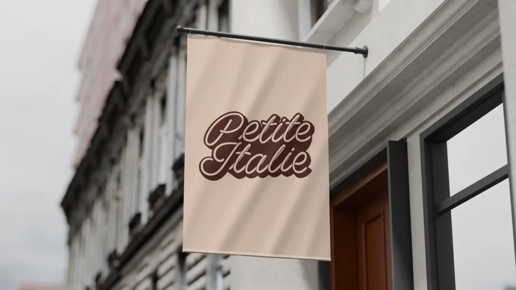 Panneau suspendu beige avec le texte 'Petite Italie' en écriture cursive marron, sur la façade d'un bâtiment urbain.