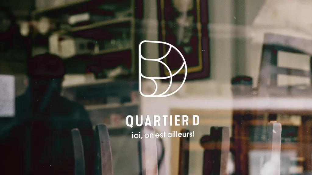 Logo 'Quartier D' blanc sur une vitrine, avec la phrase 'ici, on est ailleurs !', et un intérieur de café flou en arrière-plan.