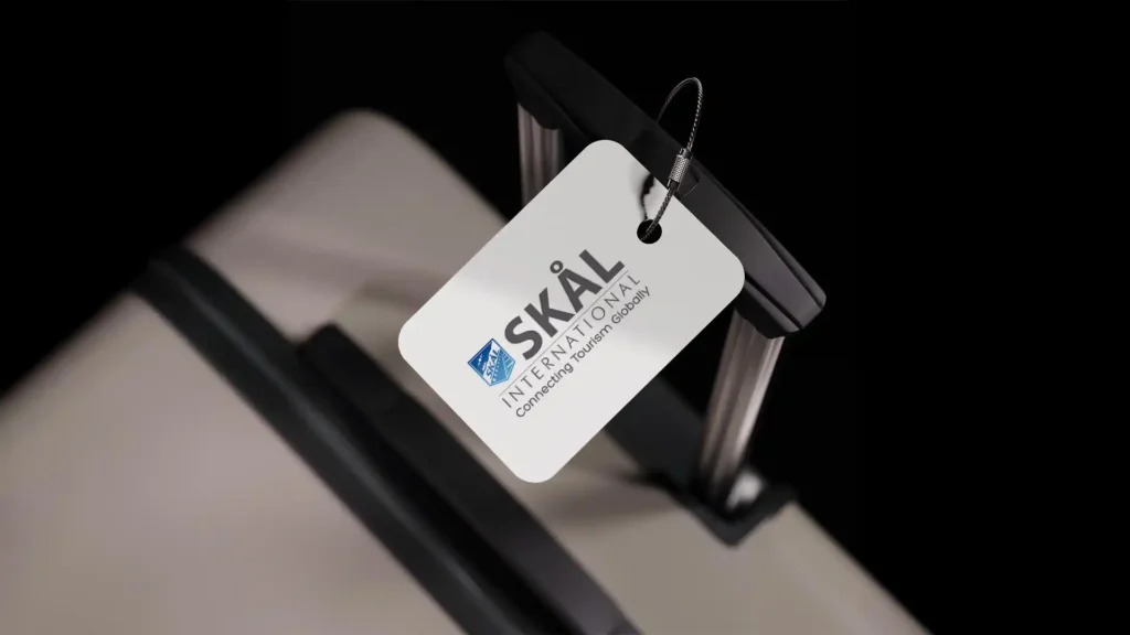 Étiquette blanche avec le logo 'SKÅL International' accrochée à une valise grise sur fond noir.